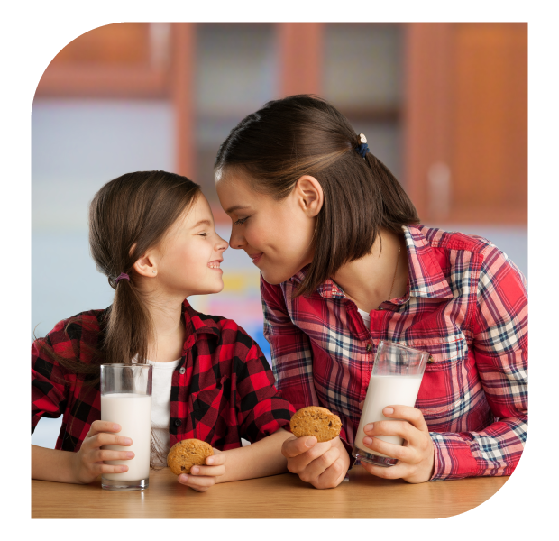Dos personas con camisas de cuadros sentadas en una cocina, cada una sosteniendo un vaso de leche y una galleta. Ambiente cálido y hogareño.