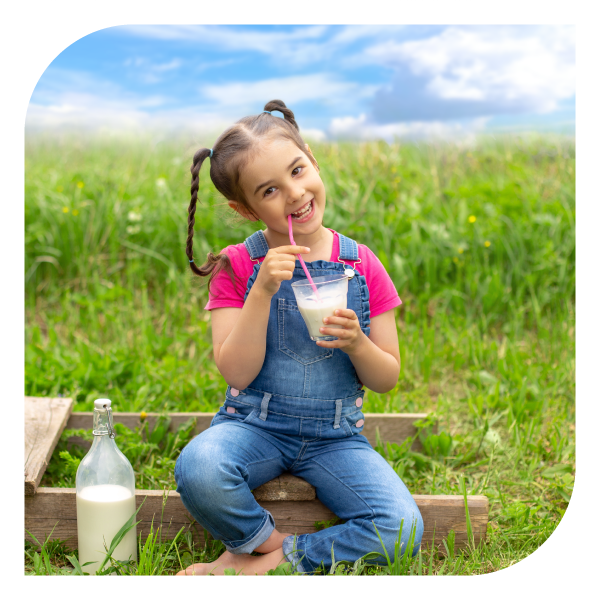 Niño con overol azul tomando leche con pajilla en un campo verde bajo cielo azul, junto a una botella de leche sobre una tabla de madera.